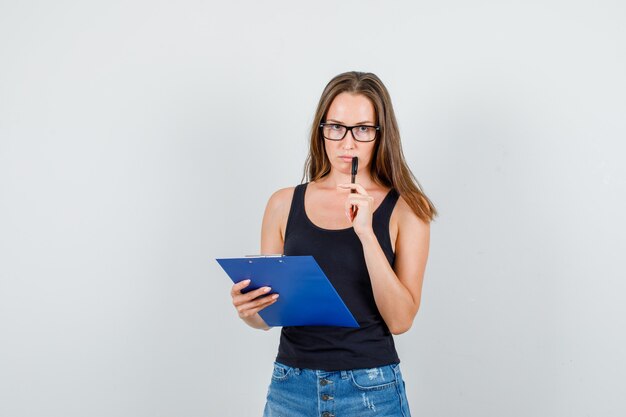 Young woman holding clipboard and pen in singlet, shorts, glasses and looking pensive