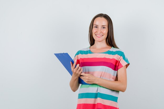 Young woman holding clipboard in t-shirt and looking happy. front view.