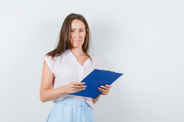 Young woman holding clipboard in t-shirt, skirt and looking sorrowful. front view.