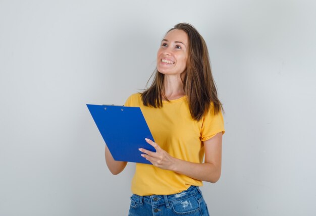 Young woman holding clipboard while looking up in t-shirt, shorts and looking cheery