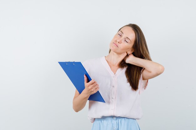 Young woman holding clipboard with hand on neck in t-shirt, skirt and looking tired , front view.