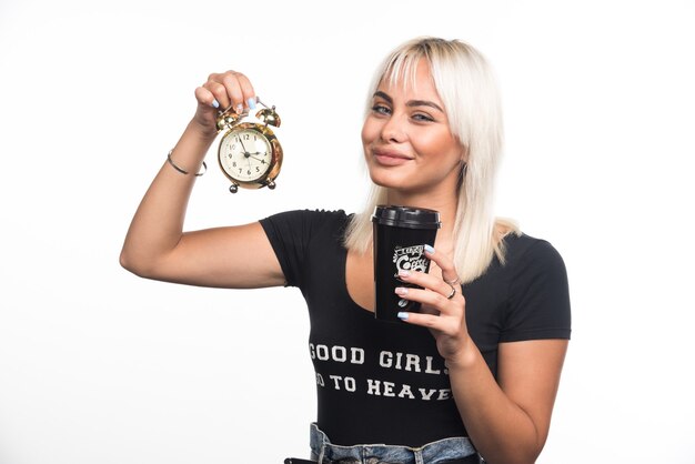 Young woman holding clock and cup of coffee on white wall.