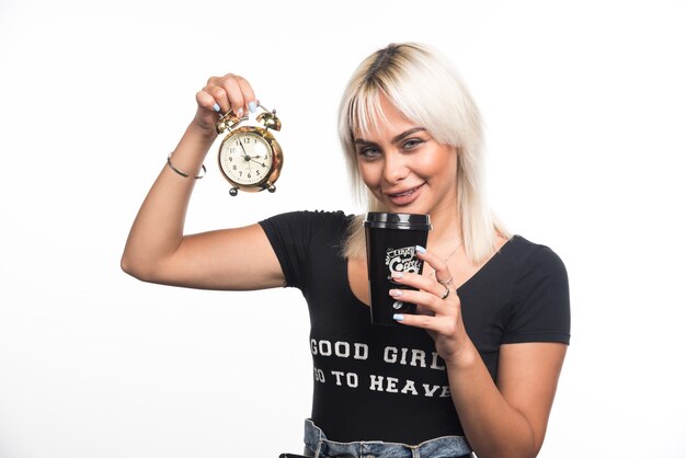 Young woman holding clock and cup of coffee on white wall.
