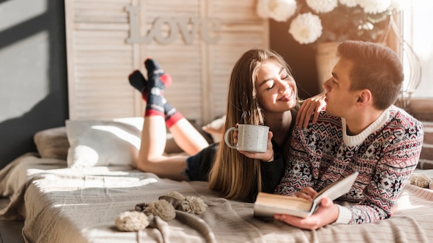 Young woman holding coffee cup in hand looking at man lying on bed