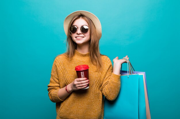 Young woman holding coffee to go and shopping bags while smiling on blue wall