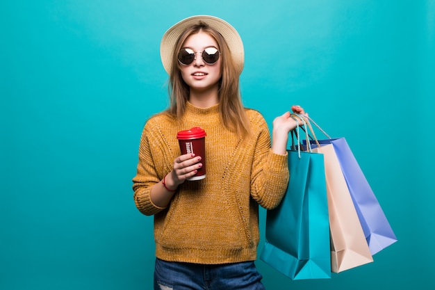 Young woman holding coffee to go and shopping bags while smiling on blue wall