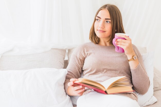 Young woman holding coffee sitting on bed holding book