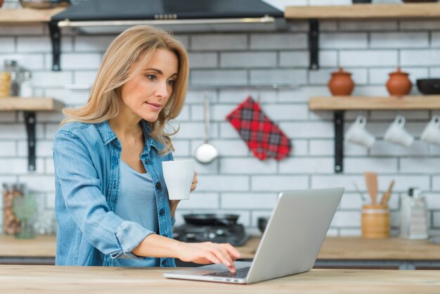 Young woman holding cup of coffee typing on laptop in the kitchen