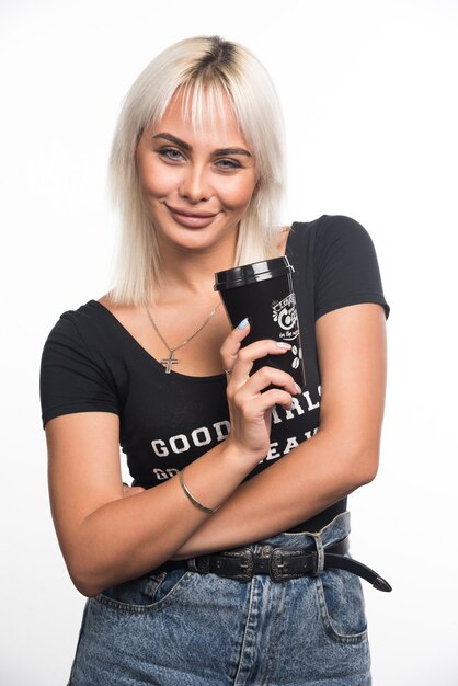 Young woman holding cup of coffee on white wall with arms crossed.