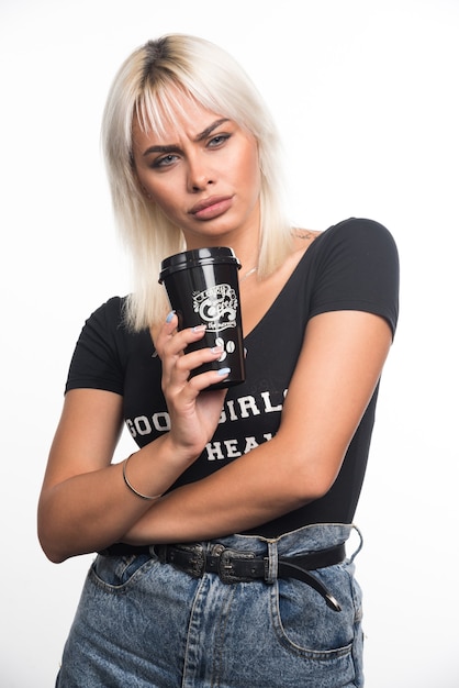 Young woman holding cup of coffee on white wall with serious expression.
