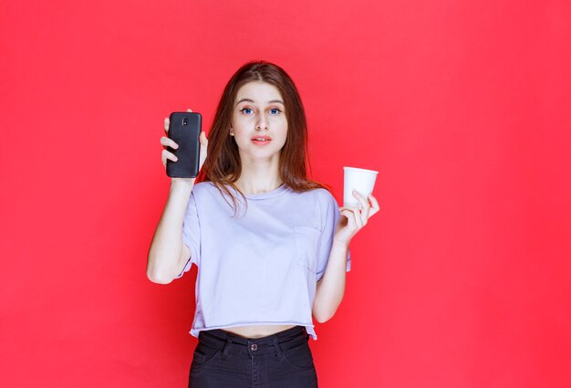 young woman holding a cup of drink and a black smartphone with a disappointed face.