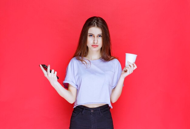 young woman holding a cup of drink and a black smartphone with a disappointed face.