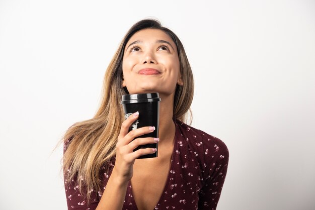 Young woman holding a cup of drink on white wall. 