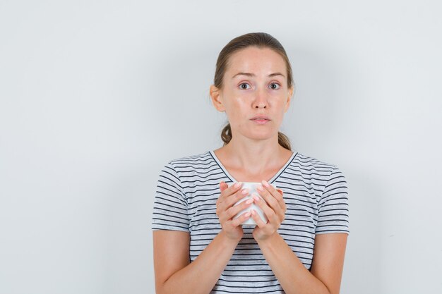 Young woman holding cup of tea in t-shirt and looking confused. front view.