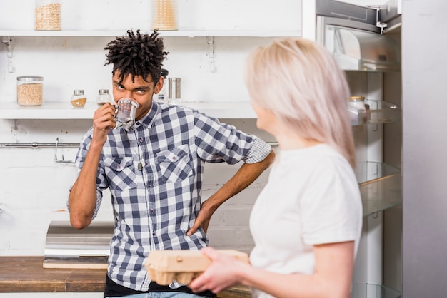 Young woman holding egg carton in hand looking at her boyfriend drinking the coffee