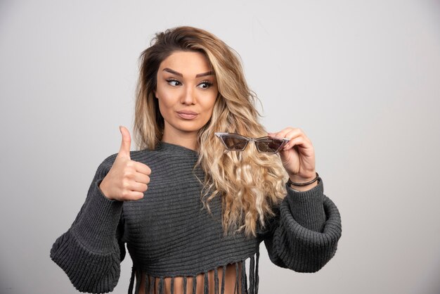 Young woman holding eyeglasses and giving thumbs up.