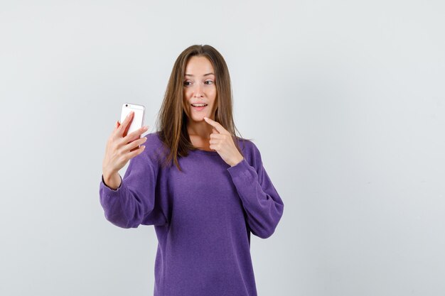 Young woman holding finger on chin while taking selfie in violet shirt front view.