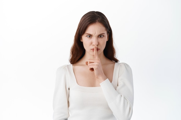Young woman holding finger on lips mouth to keep it quiet hush, standing isolated on white blank studio wall with copy space, millennial gossip girl showing shh gesture secret in silence