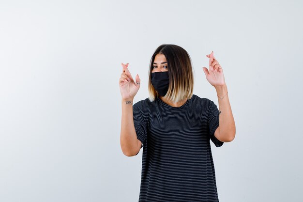 Young woman holding fingers crossed in black dress, black mask and looking happy. front view.