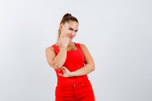 Young woman holding fist on chin in red tank top, pants and looking attractive , front view.