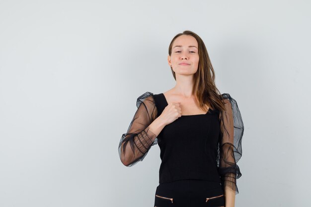 Молодая женщина держит кулак на груди в черной блузке и выглядит надменно