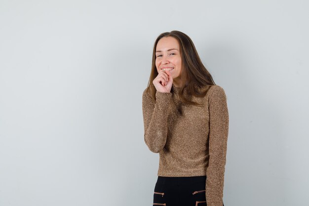 Young woman holding fist on her jaw while laughing in golden blouse and looking jolly