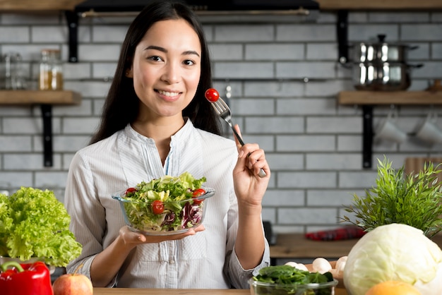 Young woman holding fork with tomato and healthy salad standing in kitchen