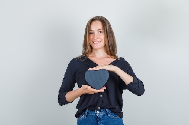 Contenitore di regalo della tenuta della giovane donna in camicia nera, pantaloncini di jeans e che sembra gioiosa