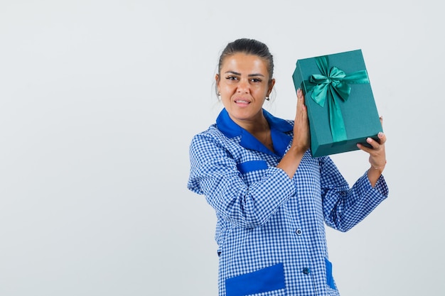 Giovane donna che tiene confezione regalo in camicia pigiama a quadretti blu e sembra carina, vista frontale.