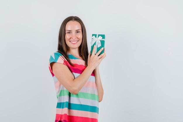Young woman holding gift box in t-shirt and looking happy. front view.