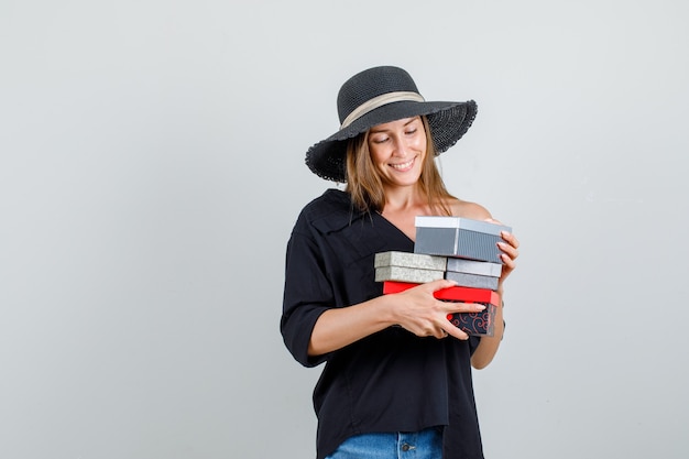 Молодая женщина держит подарочные коробки в рубашке, шортах, шляпе и выглядит весело