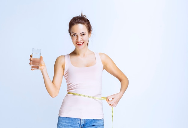 Free photo young woman holding a glass of pure water and measuring her waist size