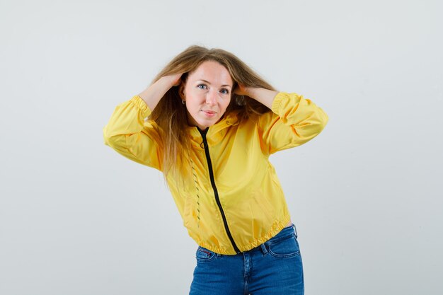 Young woman holding hair and posing at camera in yellow bomber jacket and blue jean and looking attractive. front view.