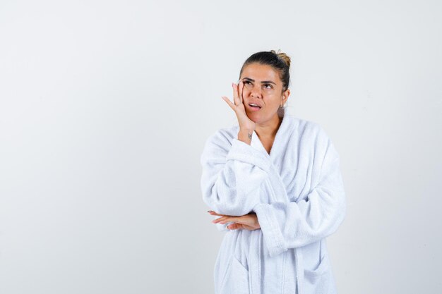 Young woman holding hand on cheek in bathrobe and looking thoughtful