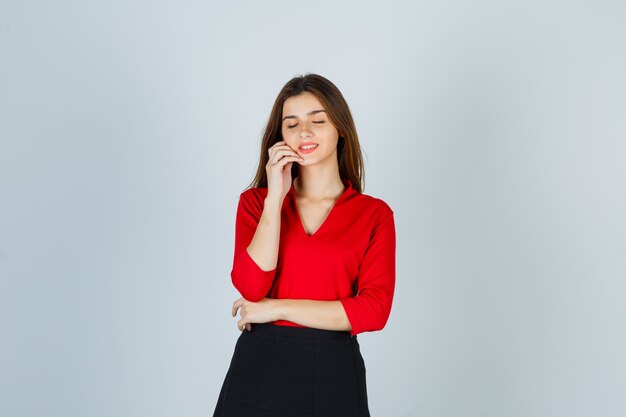 Young woman holding hand on cheek, keeping eyes closed while posing in red blouse, black skirt and looking cheerful