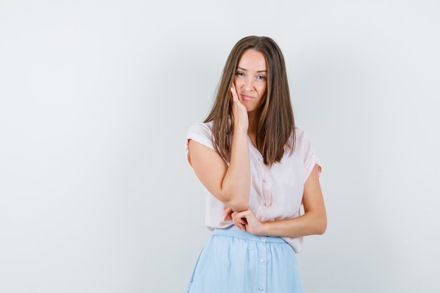 Young woman holding hand on cheek in t-shirt, skirt and looking displeased , front view.