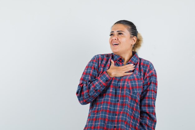 Young woman holding hand on chest in checked shirt and looking impressed. front view.