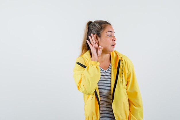 Young woman holding hand behind ear in t-shirt, jacket and looking curious , front view.