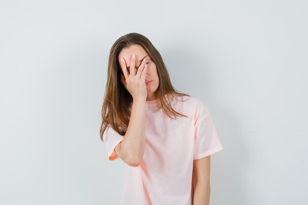 Молодая женщина держит руку на лице в розовой футболке и смотрит задумчиво.