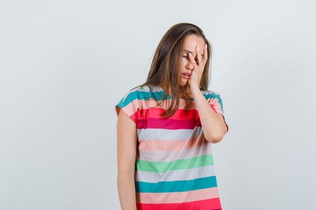 Young woman holding hand on face in t-shirt and looking annoyed , front view.