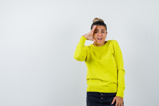 Young woman holding hand to face in yellow sweater and black pants and looking surprised