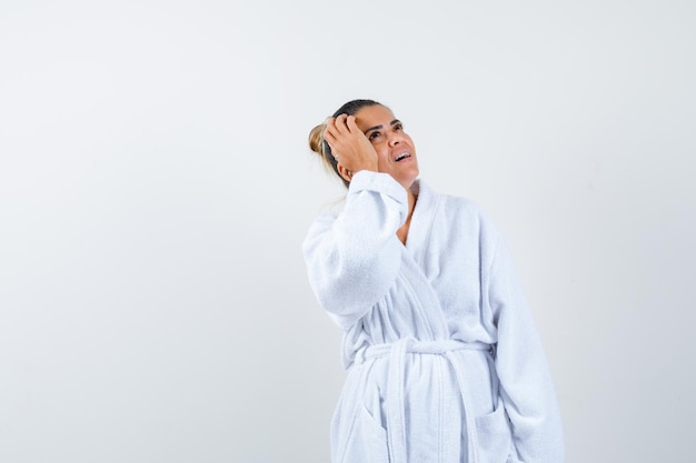 Young woman holding hand on head while looking up in bathrobe and looking thoughtful