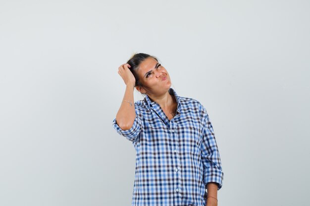 Young woman holding hand on head while thinking in checkered shirt and looking puzzled.