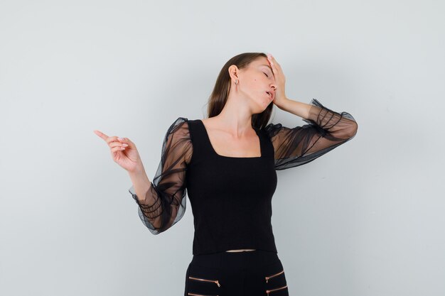 Young woman holding hand on her head while pointing aside in black blouse and looking troubled. front view.