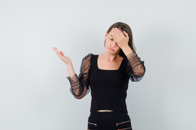 Young woman holding hand on her head while showing something at left in black blouse and looking stressful 