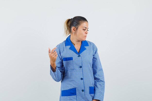 Young woman holding hand like trying to take selfie in blue gingham pajama shirt and looking pretty , front view.