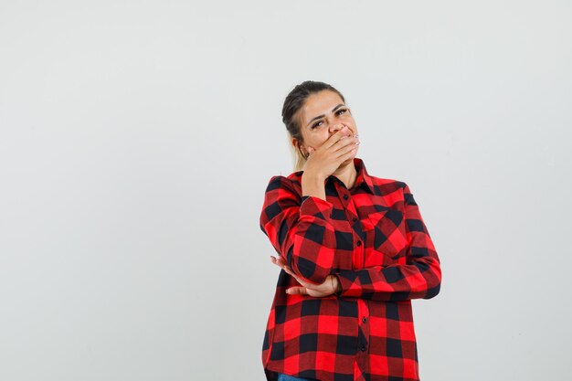 Young woman holding hand on mouth in checked shirt and looking sorry. front view.