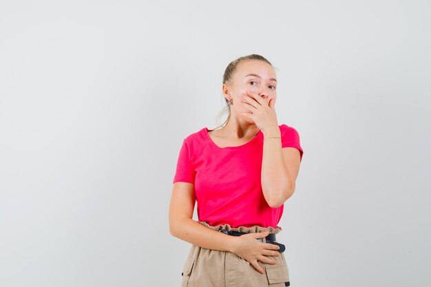 Tシャツとズボンで口に手をつないで驚いて見える若い女性