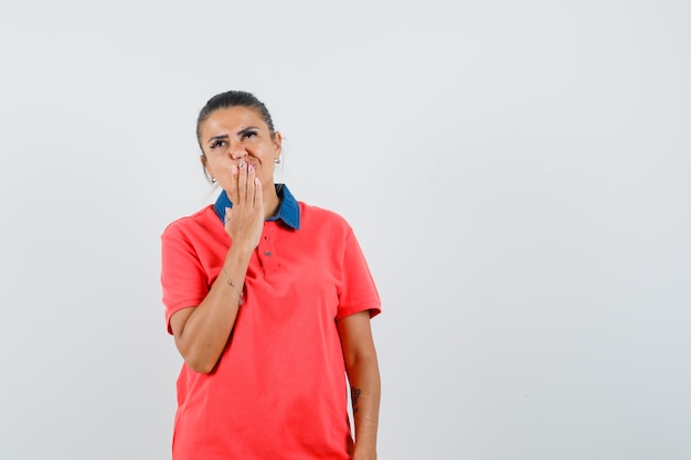 Young woman holding hand on mouth while thinking about something in red t-shirt and looking pensive , front view.