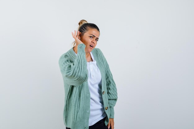 Young woman holding hand near ear to hear something in white t-shirt and mint green cardigan and looking focused